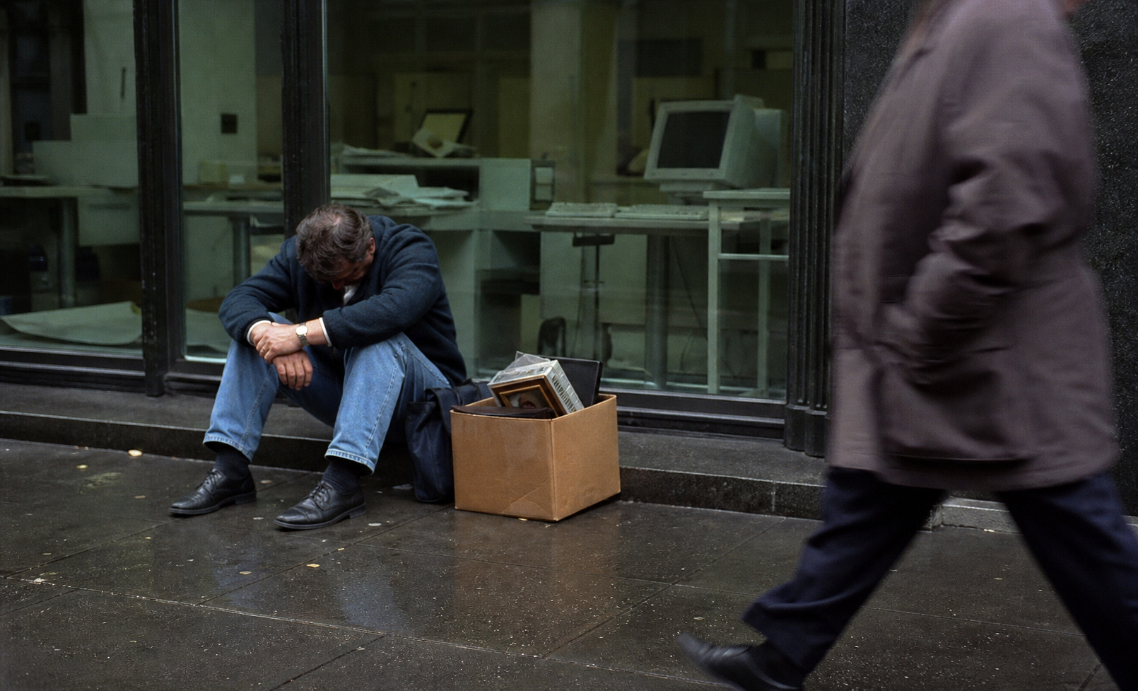 Worker sitting on a rainy sidewalk outside an emptied early 2000s office with belongings in a box.